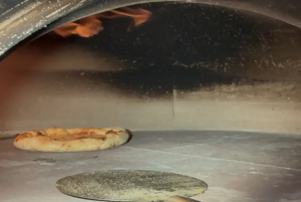 Blick in den Steinofen: offene Flamme unter der Kuppel, eine fertige Pizza auf dem Backstein, die nächste kommt auf dem Schieber herein