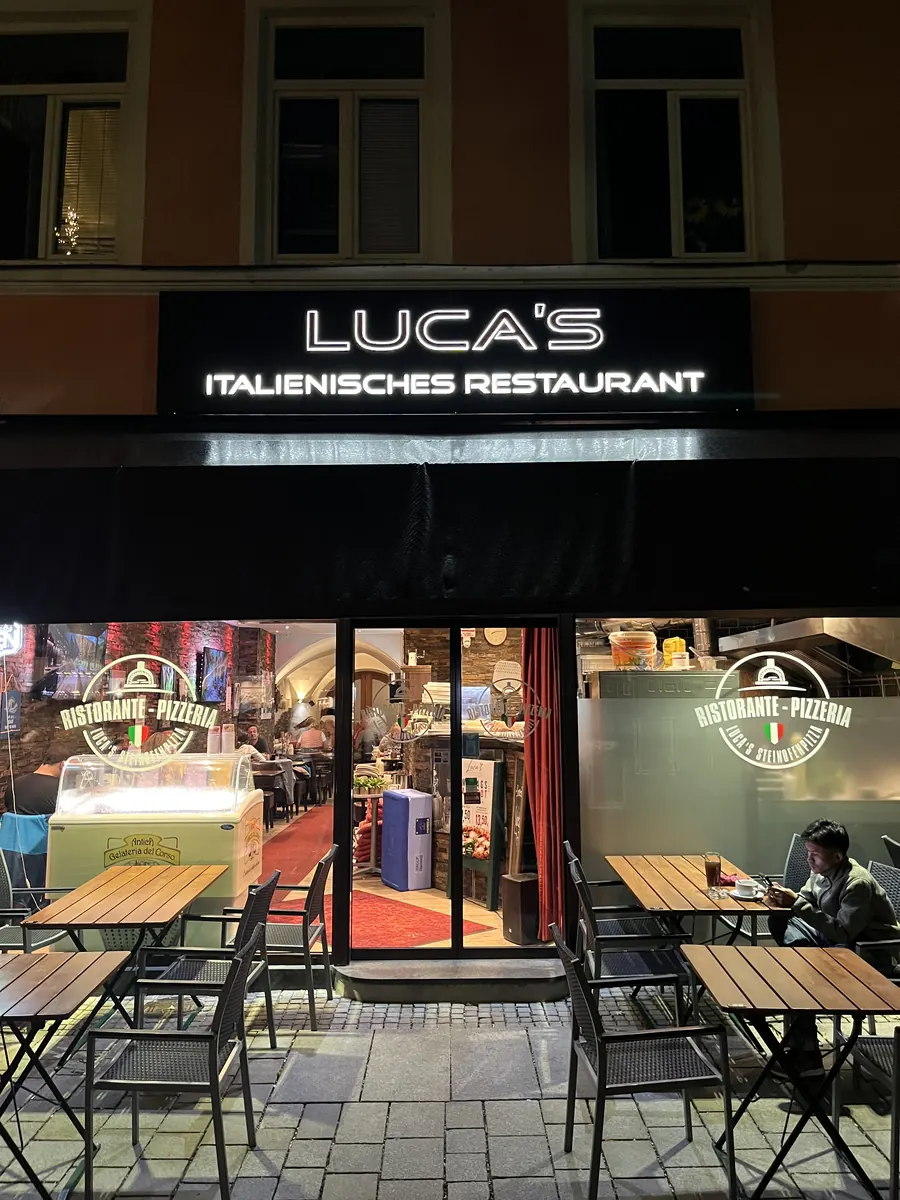Luca’s Ristorante an der Poststraße bei Nacht: beleuchteter Schriftzug, Gastgarten mit Holztischen und Blick in den Gastraum